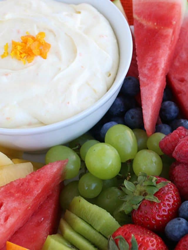 A bowl of dip made from a fruit dip recipe garnished with orange zest is surrounded by fresh fruit, including watermelon slices, green grapes, blueberries, raspberries, strawberries, kiwi, and pineapple.