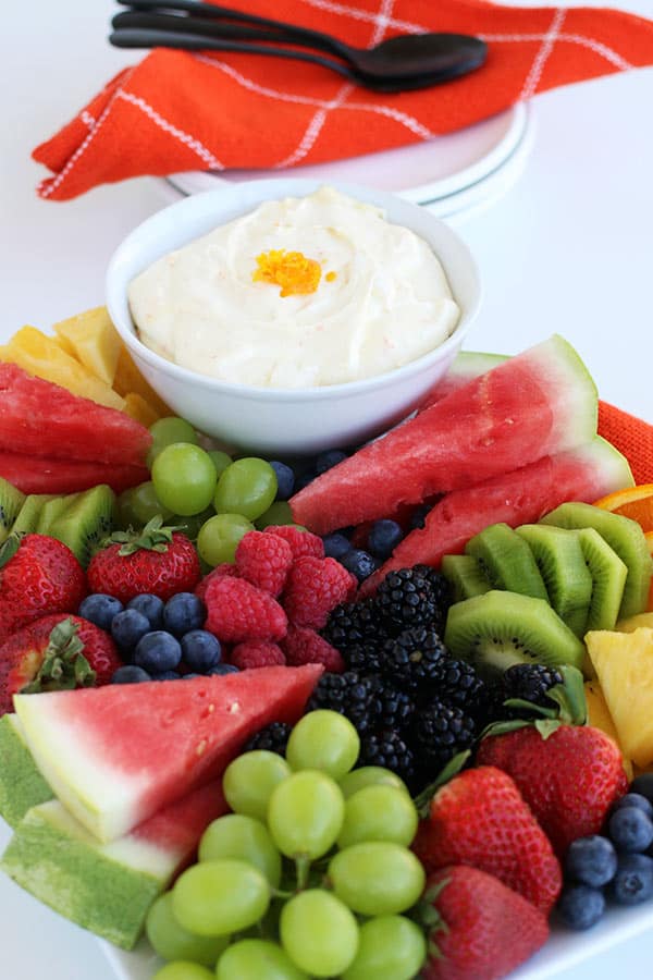A platter of assorted fresh fruits, including watermelon, strawberries, blueberries, and more, surrounds a bowl of creamy white dip—perfect for trying your favorite fruit dip recipe. Plates, utensils, and an orange napkin are in the background.