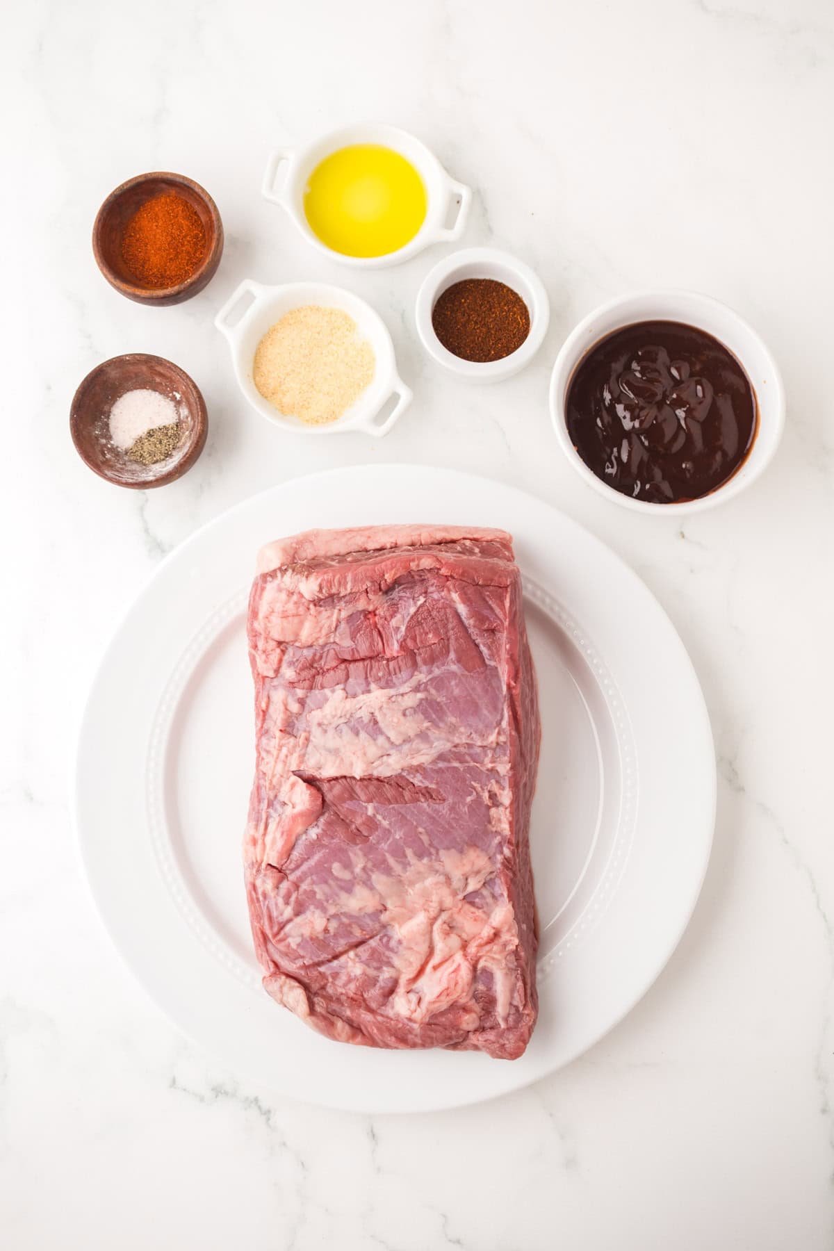 A raw beef brisket sits on a white plate, ready to become Instant Pot Brisket, surrounded by bowls of barbecue sauce, oil, brown spices, granulated garlic, and salt on a white marble surface.