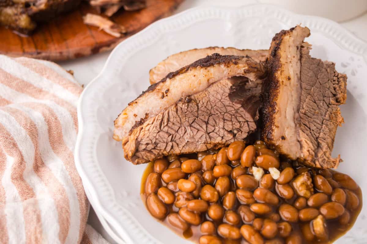 Slices of Instant Pot Brisket with a visible peppery crust are served on a white plate alongside baked beans. A striped towel and part of a wooden cutting board are visible beside the plate.
