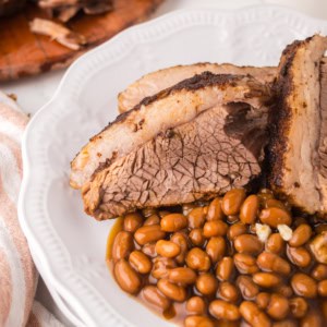 Slices of Instant Pot Brisket with a visible peppery crust are served on a white plate alongside baked beans. A striped towel and part of a wooden cutting board are visible beside the plate.