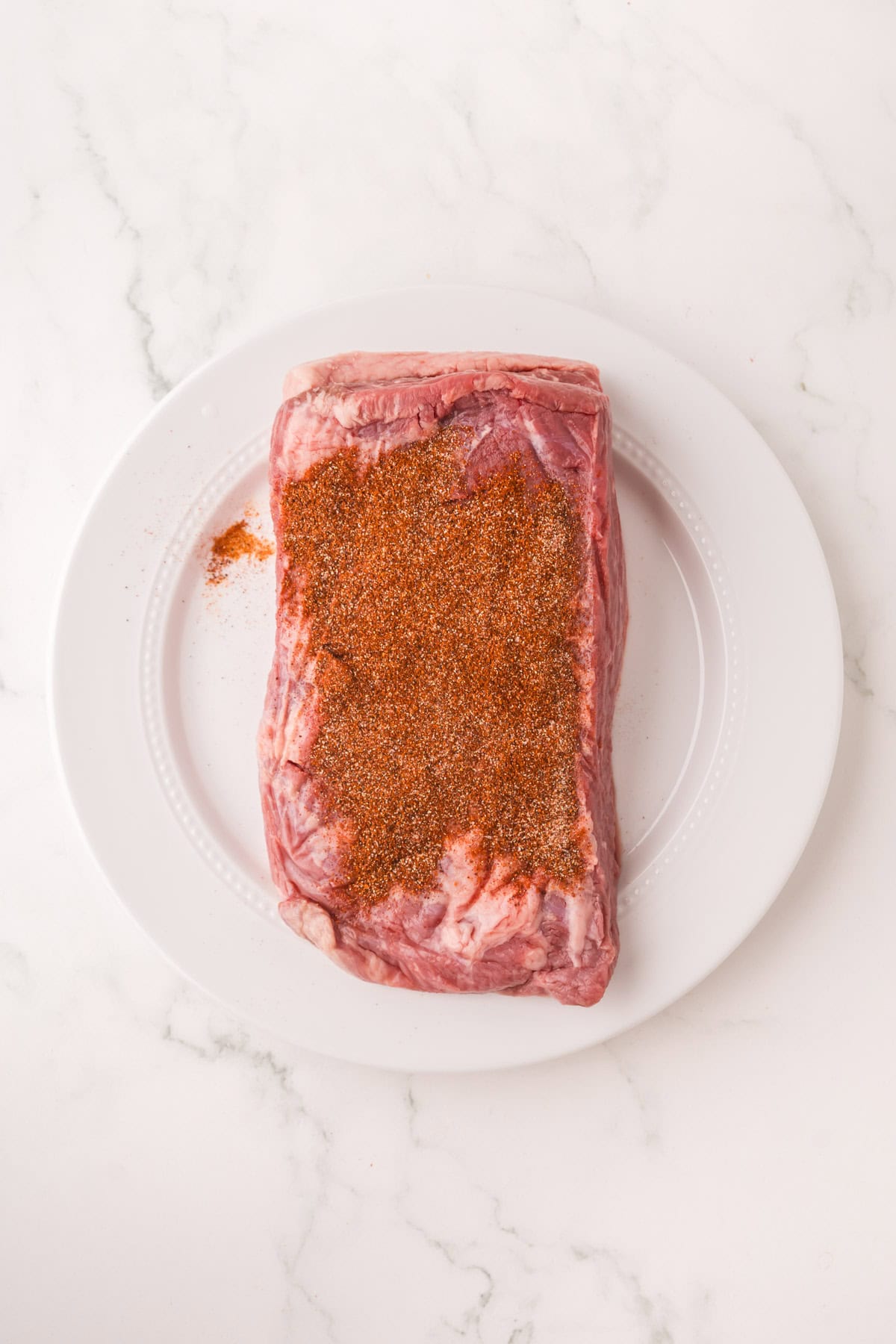 A raw cut of beef brisket on a white plate, partially covered with a reddish-brown dry spice rub, ready to become tender Instant Pot Brisket on a white marble surface.