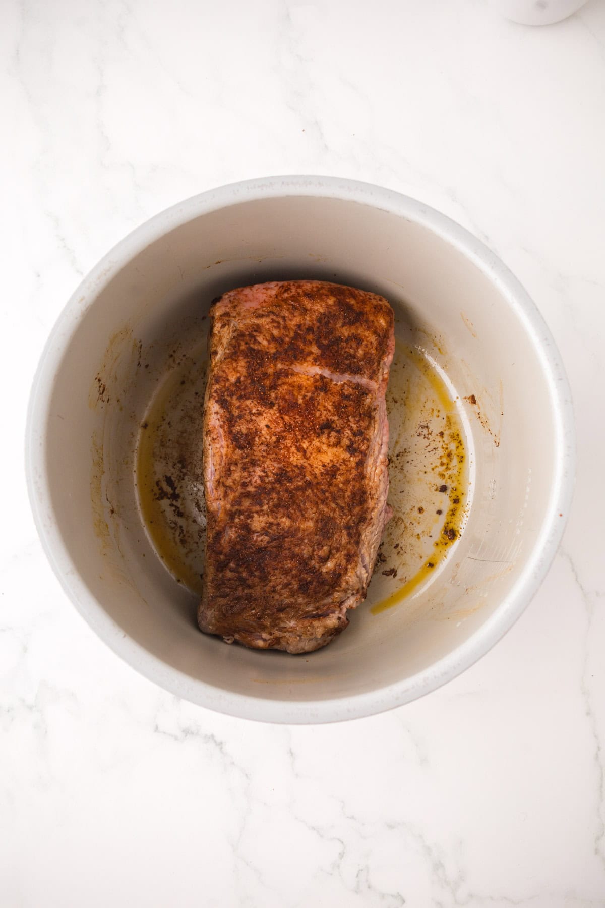 A seasoned piece of beef roast, perfect for Instant Pot Brisket, sits in the bottom of a white pot, partially browned on a marble countertop.