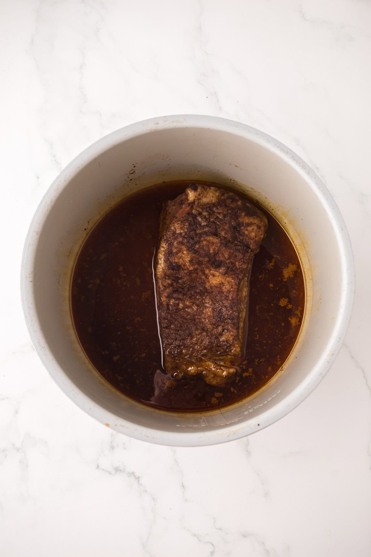 A cooked piece of seasoned meat sits in brown broth inside a round white Instant Pot Brisket slow cooker, set on a white marble surface.