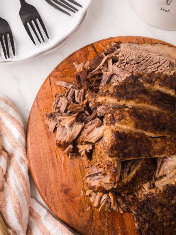 Sliced and shredded Instant Pot Brisket on a wooden cutting board, with a striped towel nearby. A plate with three black forks is partially visible in the upper left corner.