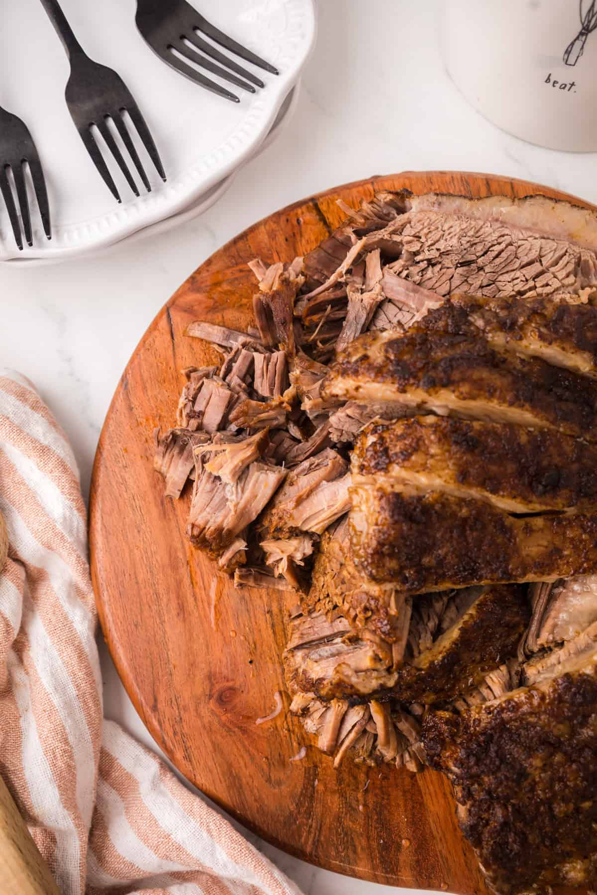 Sliced and shredded Instant Pot Brisket on a wooden cutting board, with a striped towel nearby. A plate with three black forks is partially visible in the upper left corner.
