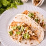 Two soft instant pot chicken tacos filled with shredded chicken, fresh cilantro, and a creamy white sauce are served on a white plate, with fresh cilantro leaves visible in the background.