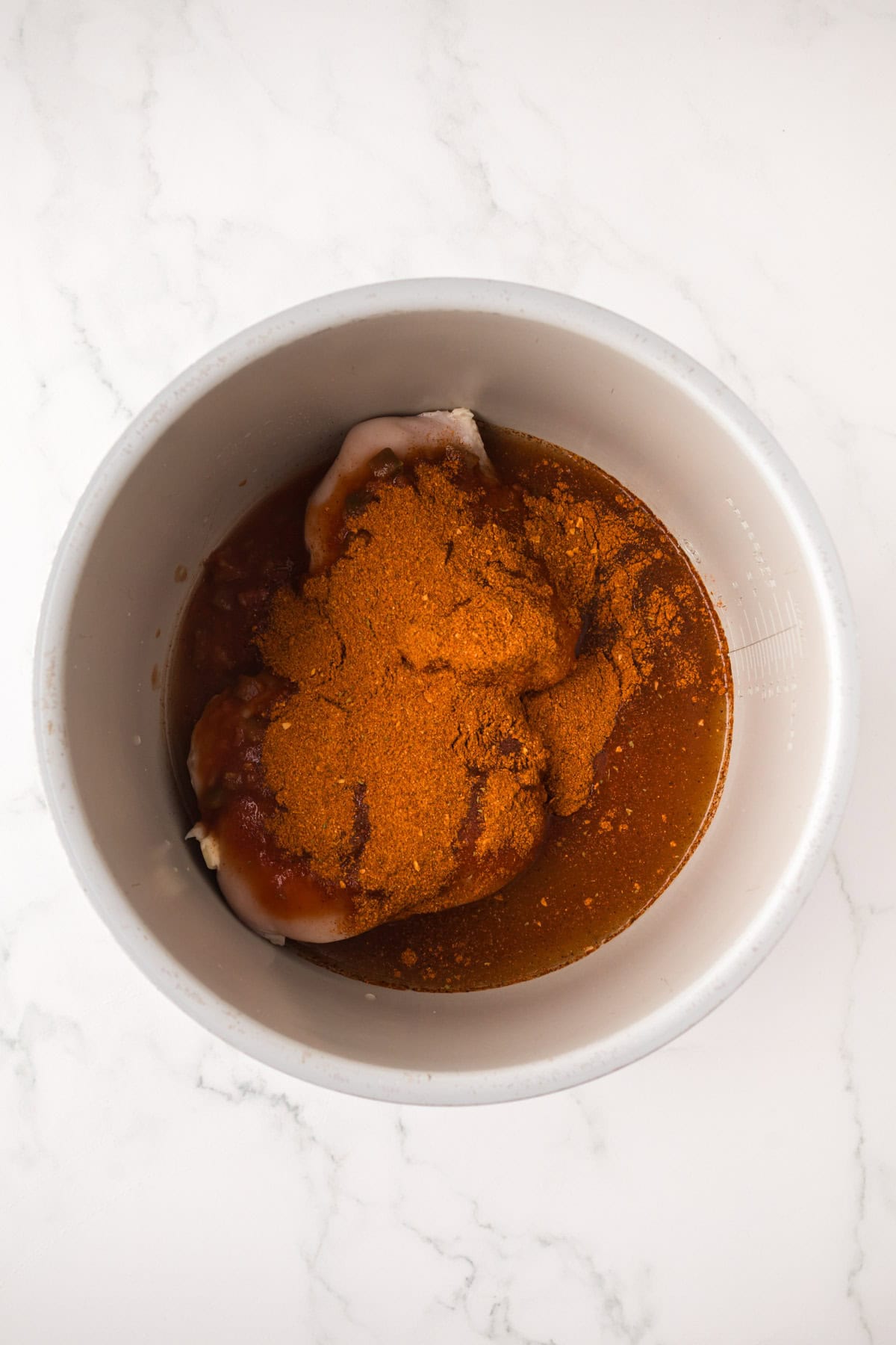Raw chicken breasts in a white bowl, topped with a reddish-brown spice mix and marinade—perfect for making flavorful Instant Pot chicken tacos—set on a white marble surface.