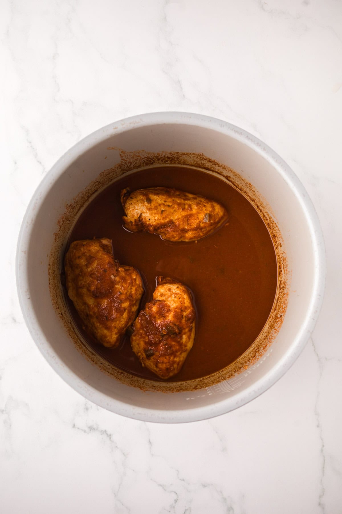 Three cooked chicken breasts in a round white pot, covered in a reddish-brown sauce, are perfect for making instant pot chicken tacos and sit on a white marble surface.