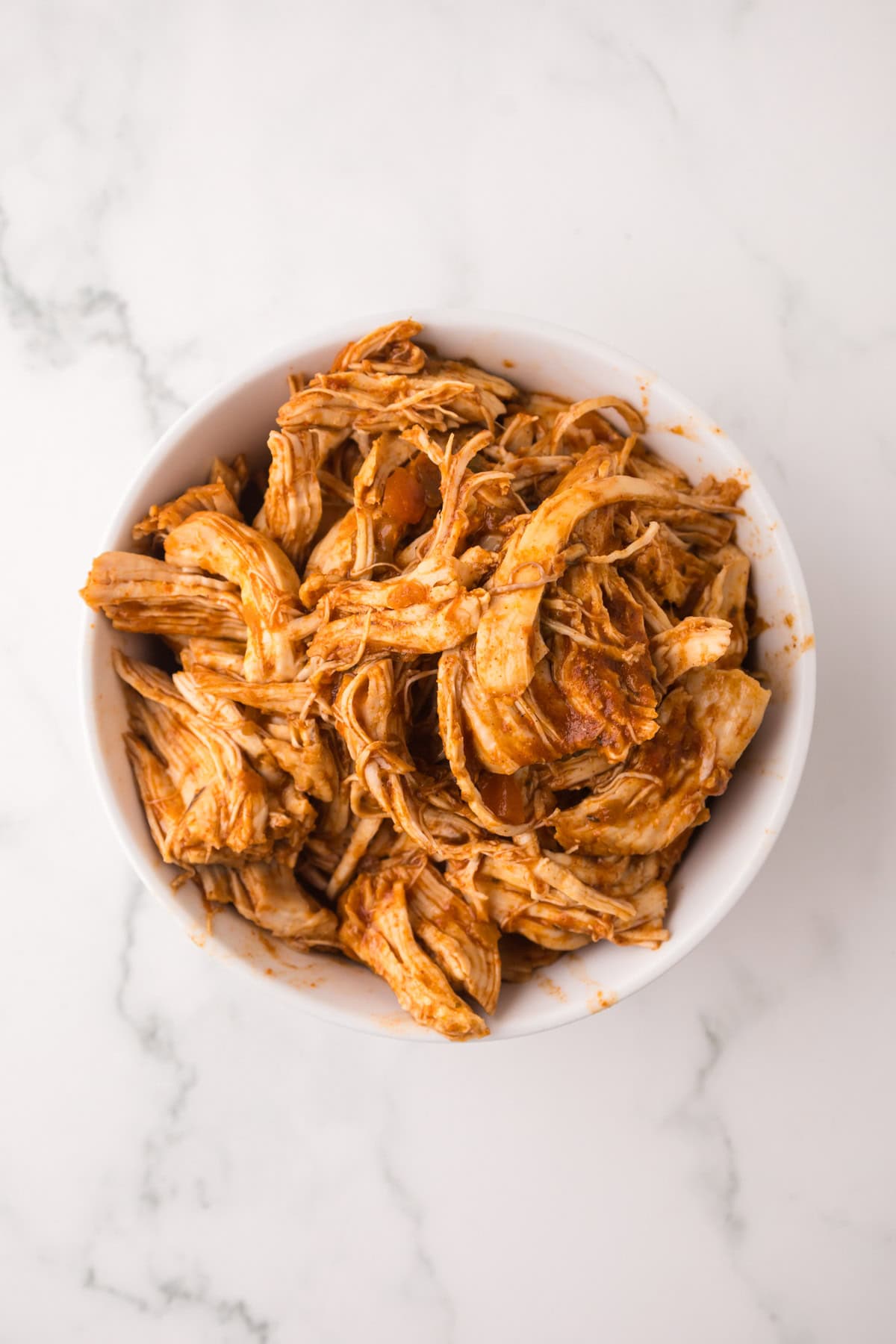 A white bowl filled with shredded, seasoned Instant Pot chicken tacos sits on a white marble surface.
