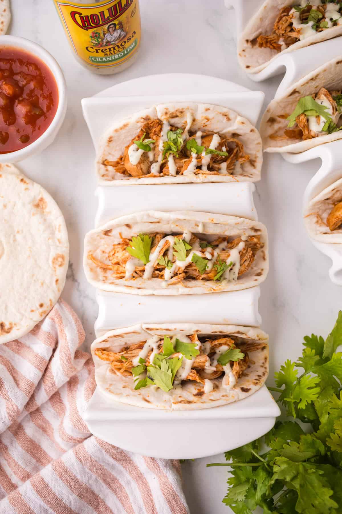 Three soft instant pot chicken tacos filled with shredded chicken, cheese, and fresh cilantro are arranged in a white taco holder on a marble surface. Nearby are a tortilla, salsa, Cholula hot sauce, a striped towel, and fresh cilantro.