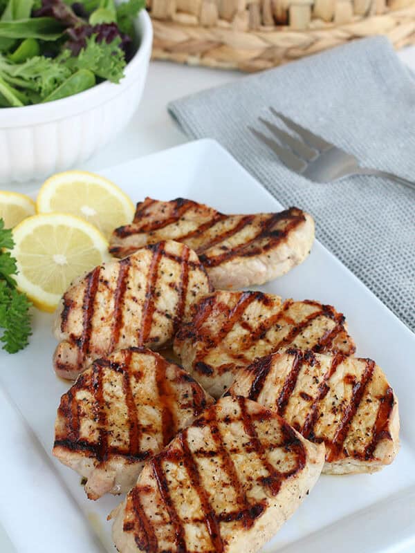 Lemon pepper porkchops with char marks on a white rectangular plate, garnished with lemon slices and parsley, alongside a bowl of mixed greens and a fork for a fresh, flavorful meal.