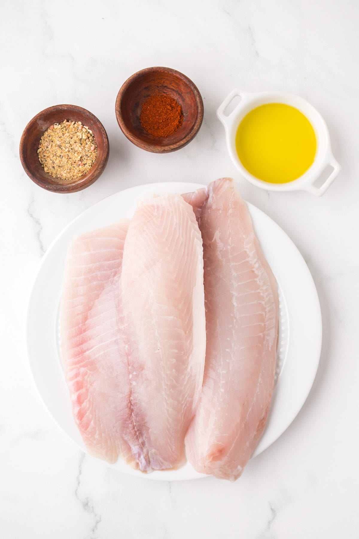 Three raw fish fillets on a white plate, ideal for pan seared tilapia, are arranged next to small bowls of yellow oil, mixed seasoning blend, and red spice on a white marble surface.