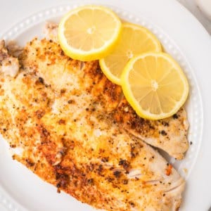 A white plate with a cooked, seasoned pan seared tilapia fillet topped with three overlapping lemon slices, set on a light surface with part of a yellow cloth visible in the corner.
