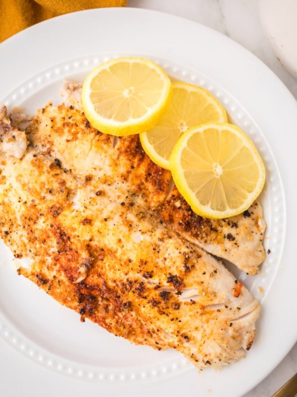 A white plate with a cooked, seasoned pan seared tilapia fillet topped with three overlapping lemon slices, set on a light surface with part of a yellow cloth visible in the corner.
