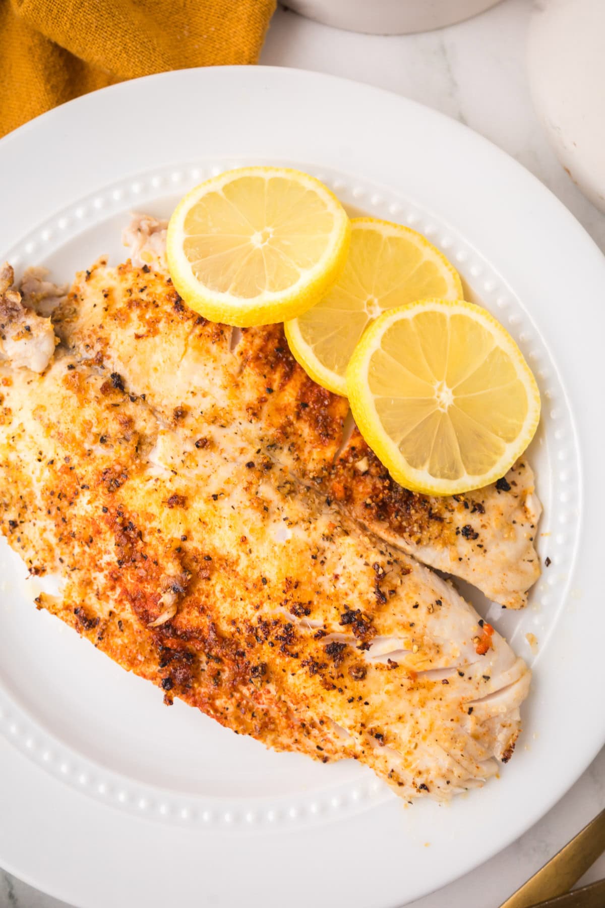 A white plate with a cooked, seasoned pan seared tilapia fillet topped with three overlapping lemon slices, set on a light surface with part of a yellow cloth visible in the corner.