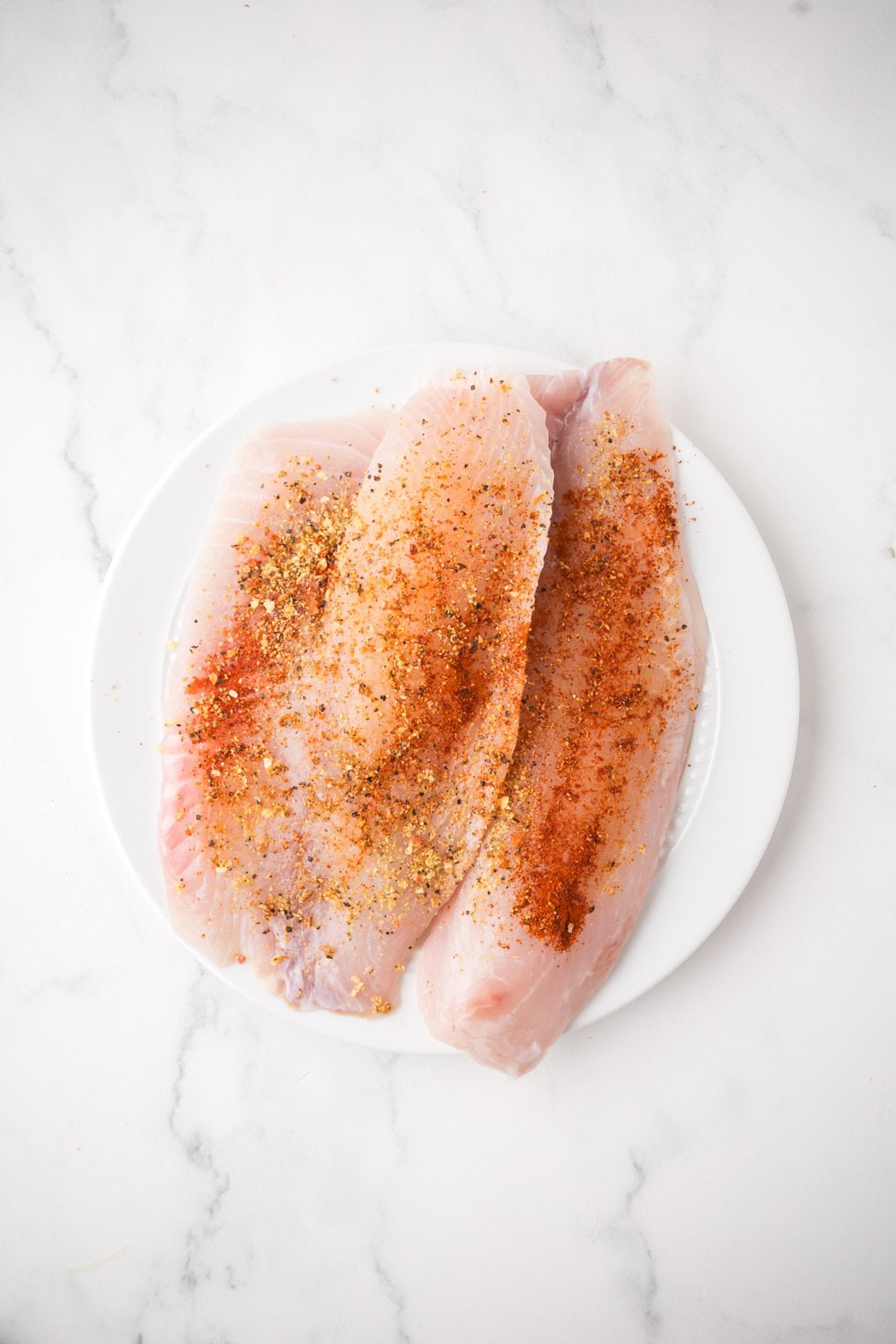 Two raw pan seared tilapia fillets on a white plate, sprinkled with a blend of red and yellow spices, are set on a white marble surface.