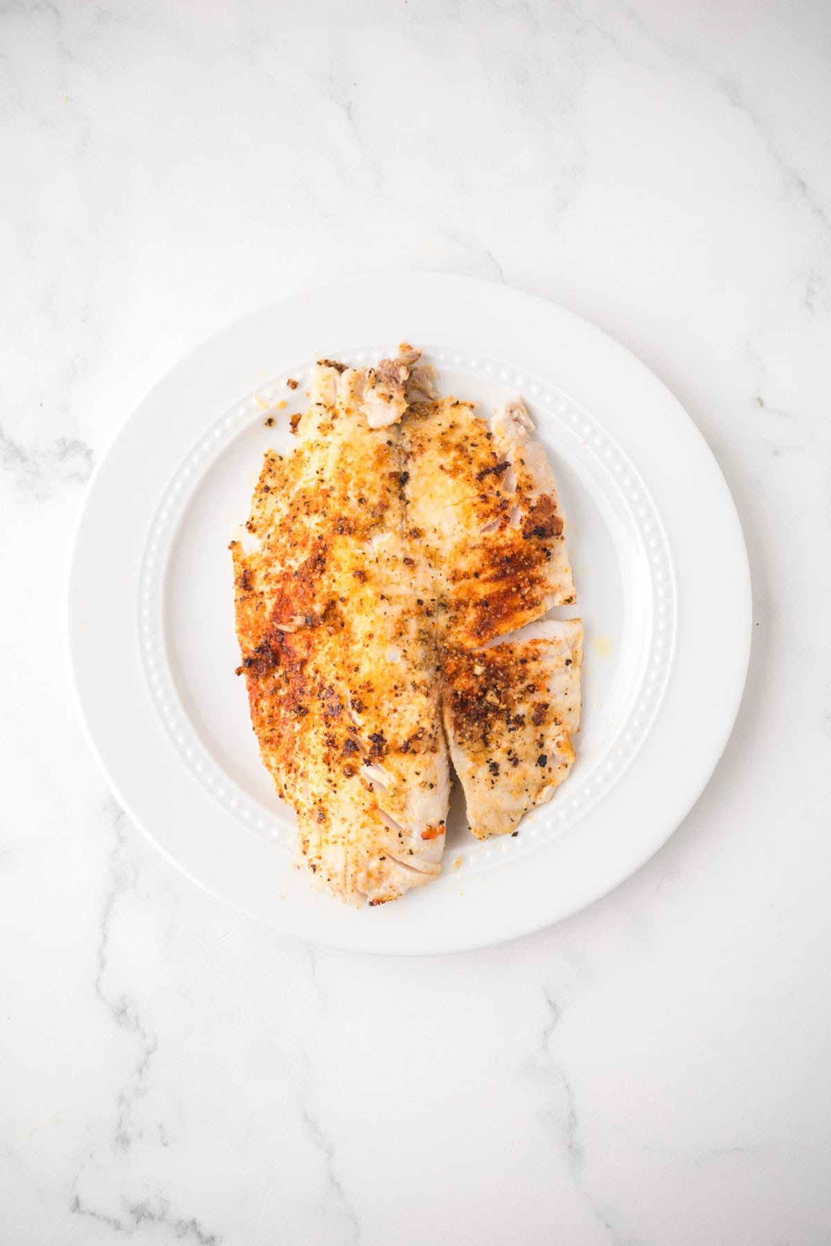 Two pieces of seasoned, pan seared tilapia fillets on a white plate, placed on a white marble surface.