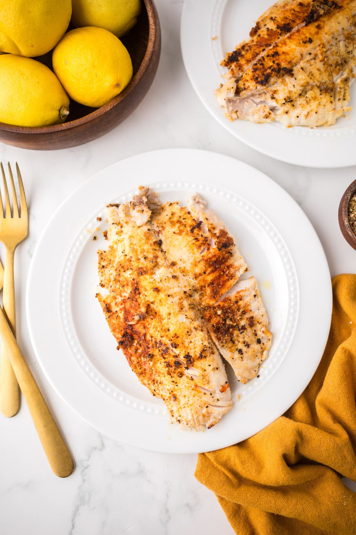 A white plate with two pan seared tilapia fillets sits next to a bowl of lemons, gold utensils, a mustard napkin, and a small bowl of spices on a white marble surface.