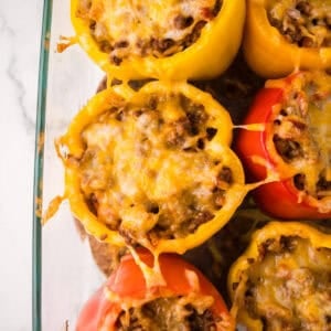 A glass baking dish filled with stuffed peppers with ground beef and melted cheese, featuring vibrant yellow and red bell peppers, viewed from above on a white surface.