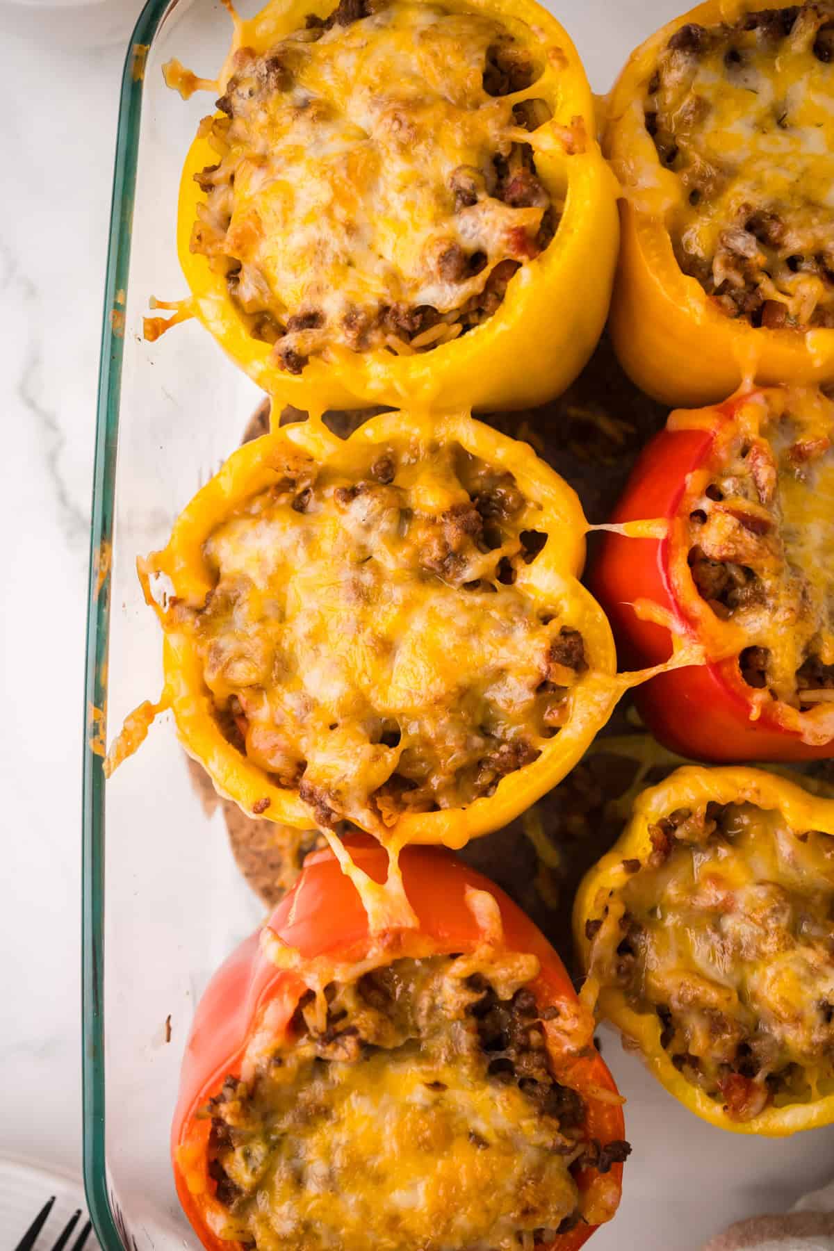 A glass baking dish filled with stuffed peppers with ground beef and melted cheese, featuring vibrant yellow and red bell peppers, viewed from above on a white surface.