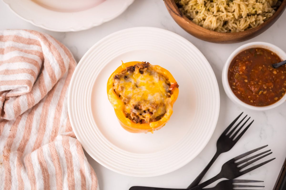 A yellow stuffed pepper with ground beef, topped with melted cheese, sits on a white plate next to rice, a bowl of salsa, a striped towel, and black utensils on a marble surface.