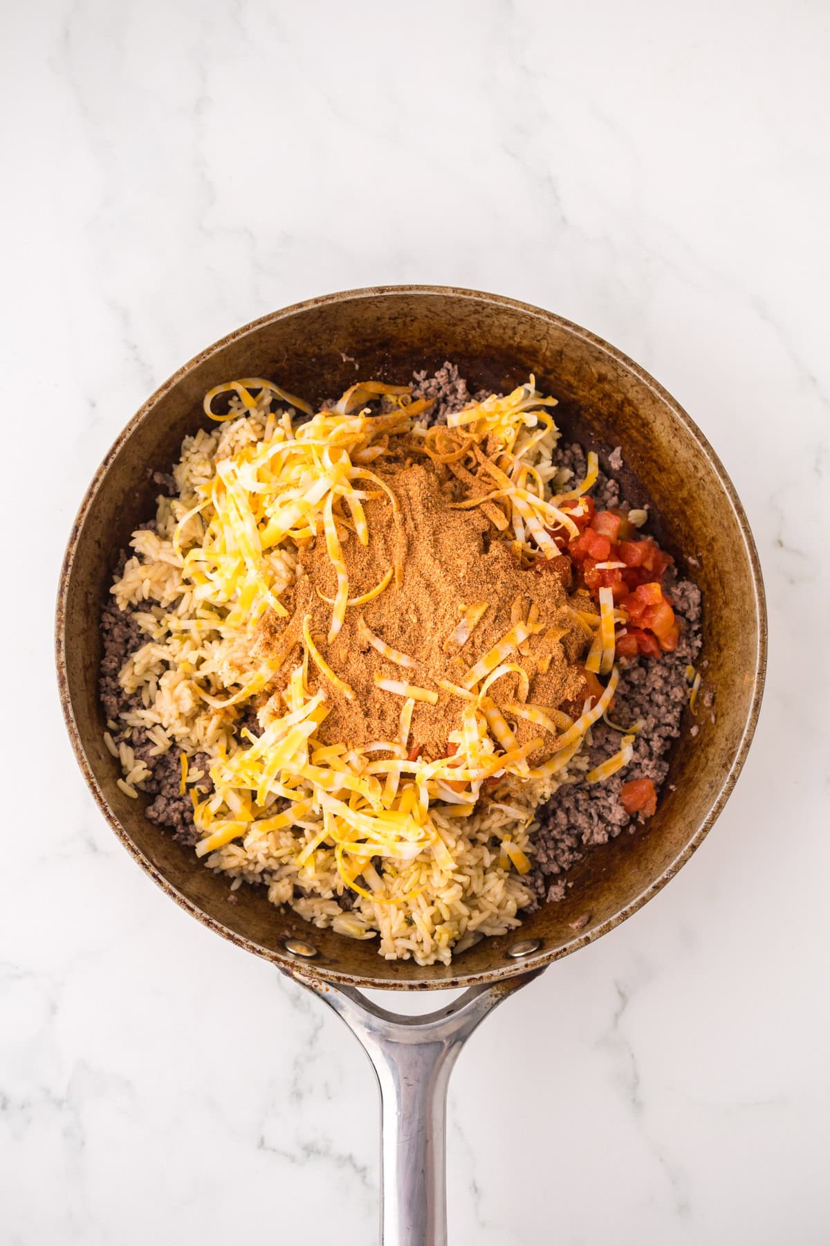 A skillet on a white marble surface contains the filling for stuffed peppers with ground beef—rice, shredded cheese, diced tomatoes, and taco seasoning all layered but not yet mixed together.