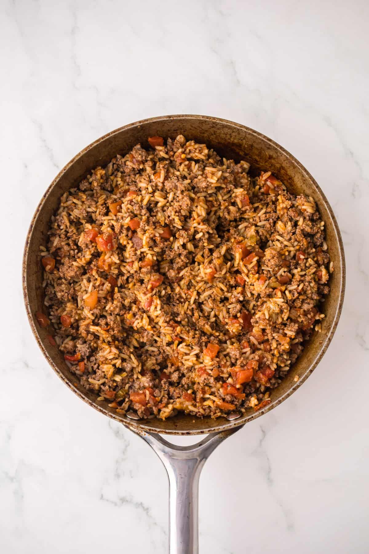 A skillet filled with a mixture reminiscent of stuffed peppers with ground beef, rice, tomatoes, and spices sits on a white marble surface.