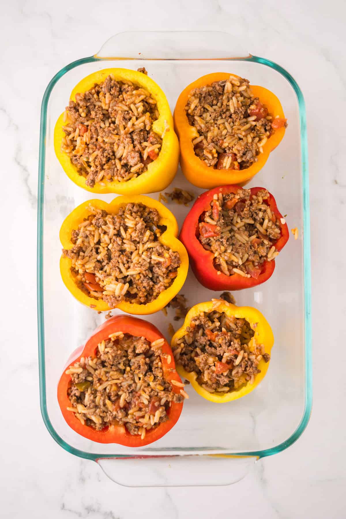 Six halved bell peppers, red and yellow, are stuffed with a savory mixture of rice, tomatoes, and ground beef, arranged in a glass baking dish on a white marble surface—a classic recipe for stuffed peppers with ground beef.