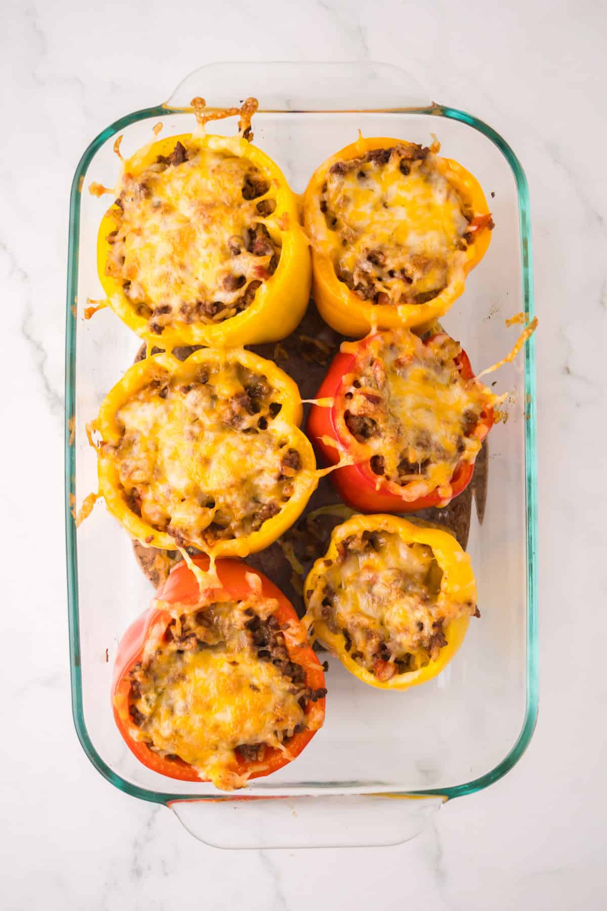 A glass baking dish with seven stuffed peppers with ground beef and rice, topped with melted cheese. The red and yellow peppers sit on a white marble surface, their savory filling peeking beneath a golden cheese layer.
