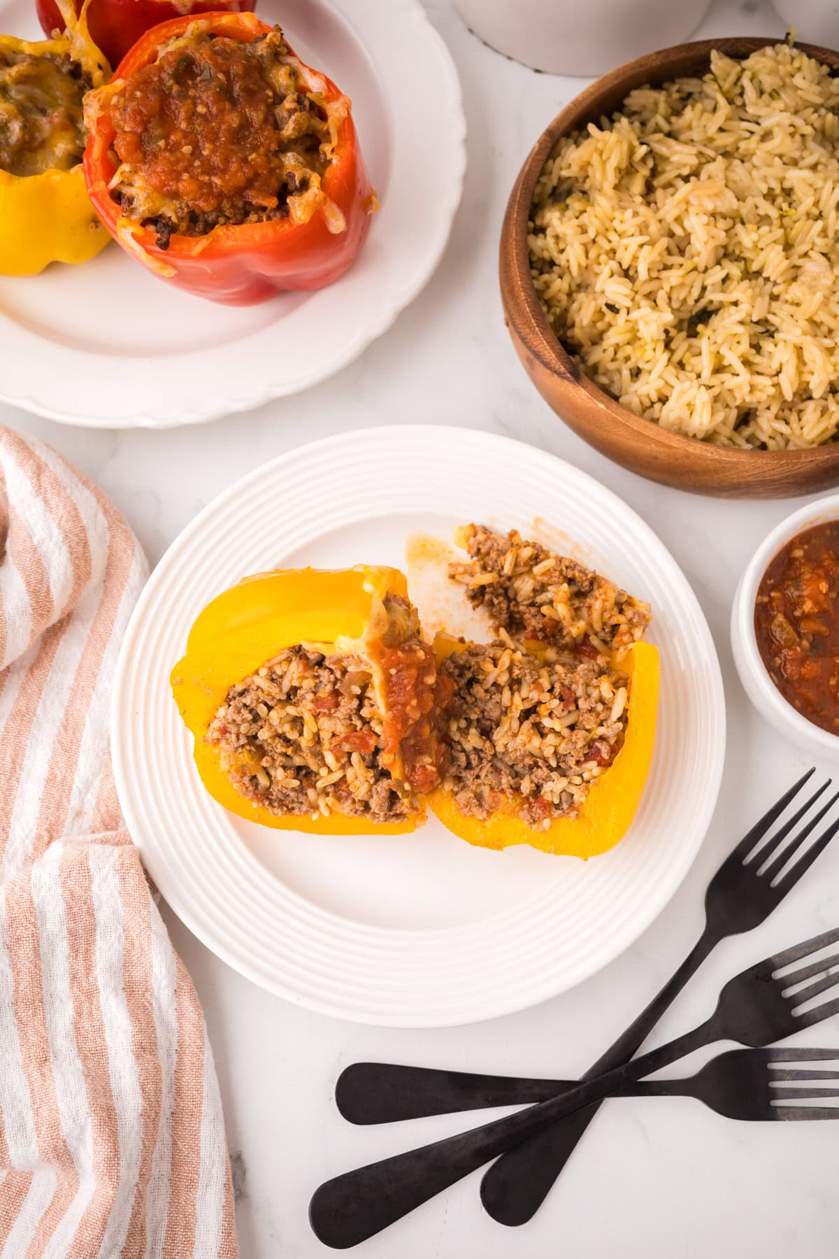 A white plate holds a yellow bell pepper stuffed with a savory rice and ground meat mixture, cut in half. Black forks, a striped cloth, a bowl of rice, salsa, and more stuffed peppers with ground beef sit nearby.