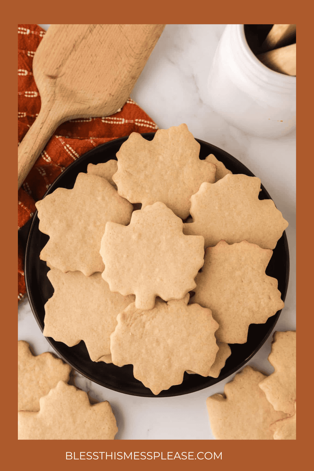 A black plate filled with maple leaf-shaped sugar cookies sits on a white surface, next to a wooden spatula, an orange checkered cloth, and a white jar—perfect inspiration for your fall cookie recipes.