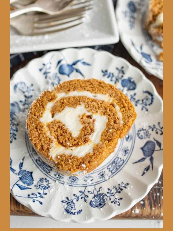 A slice of pumpkin roll cake with cream cheese filling, a classic among fall desserts, sits on a decorative blue and white plate; a fork is nearby. The plate rests on a wooden surface with another plate partly visible showing off cozy fall desserts.