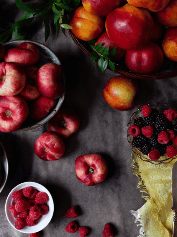 A flat lay of various fresh fruits, including nectarines, peaches, raspberries, and blackberries on a dark surface, with a yellow cloth and a BLESSTHISMESSPLEASE.COM label at the bottom—perfect inspiration for learning how to preserve plums.
