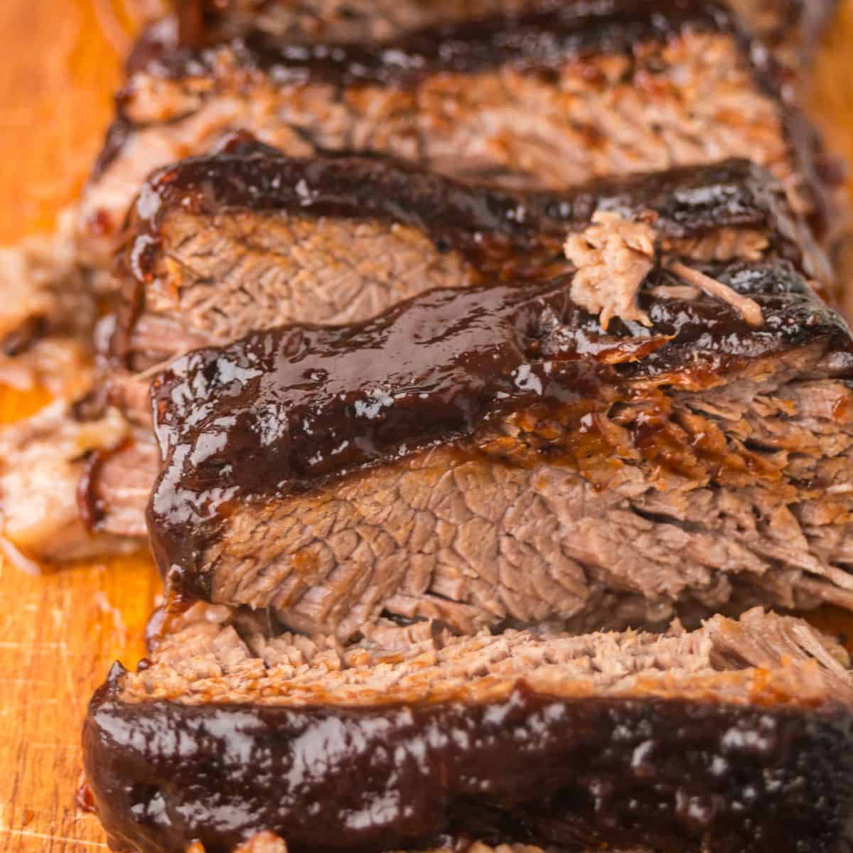 Close-up of several thick slices of slow cooker brisket with a dark, glossy layer of barbecue sauce on top, displayed on a wooden cutting board.