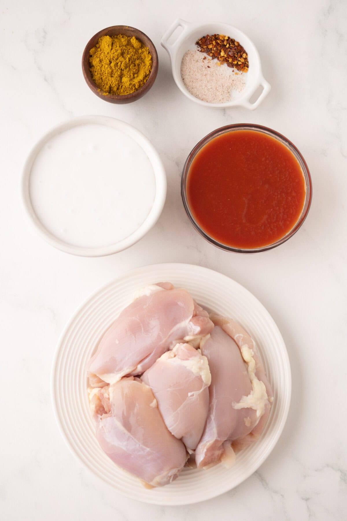 A plate with raw chicken thighs, a bowl of coconut milk, tomato sauce, curry powder, and a dish of salt and red chili flakes arranged on a white surface—these are perfect ingredients for making delicious chicken korma.