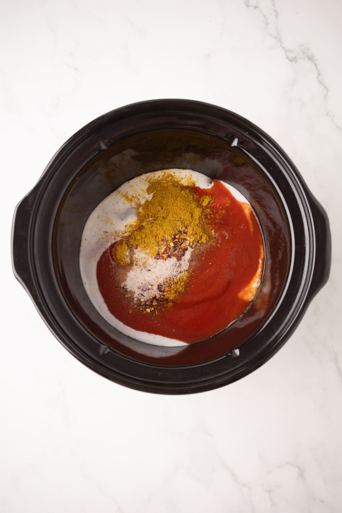 A black slow cooker containing chicken korma with a mixture of red sauce, white cream, and yellow powdered spices sits on a white marble surface.