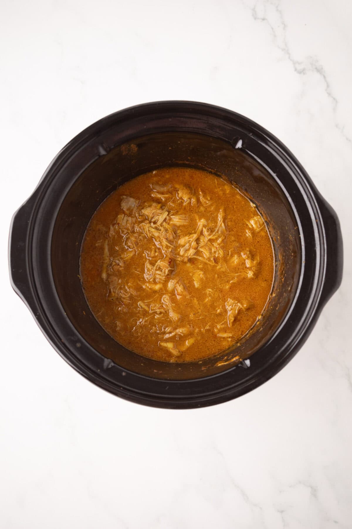 A crockpot filled with shredded chicken korma in a rich brown sauce, sitting on a white marble surface.