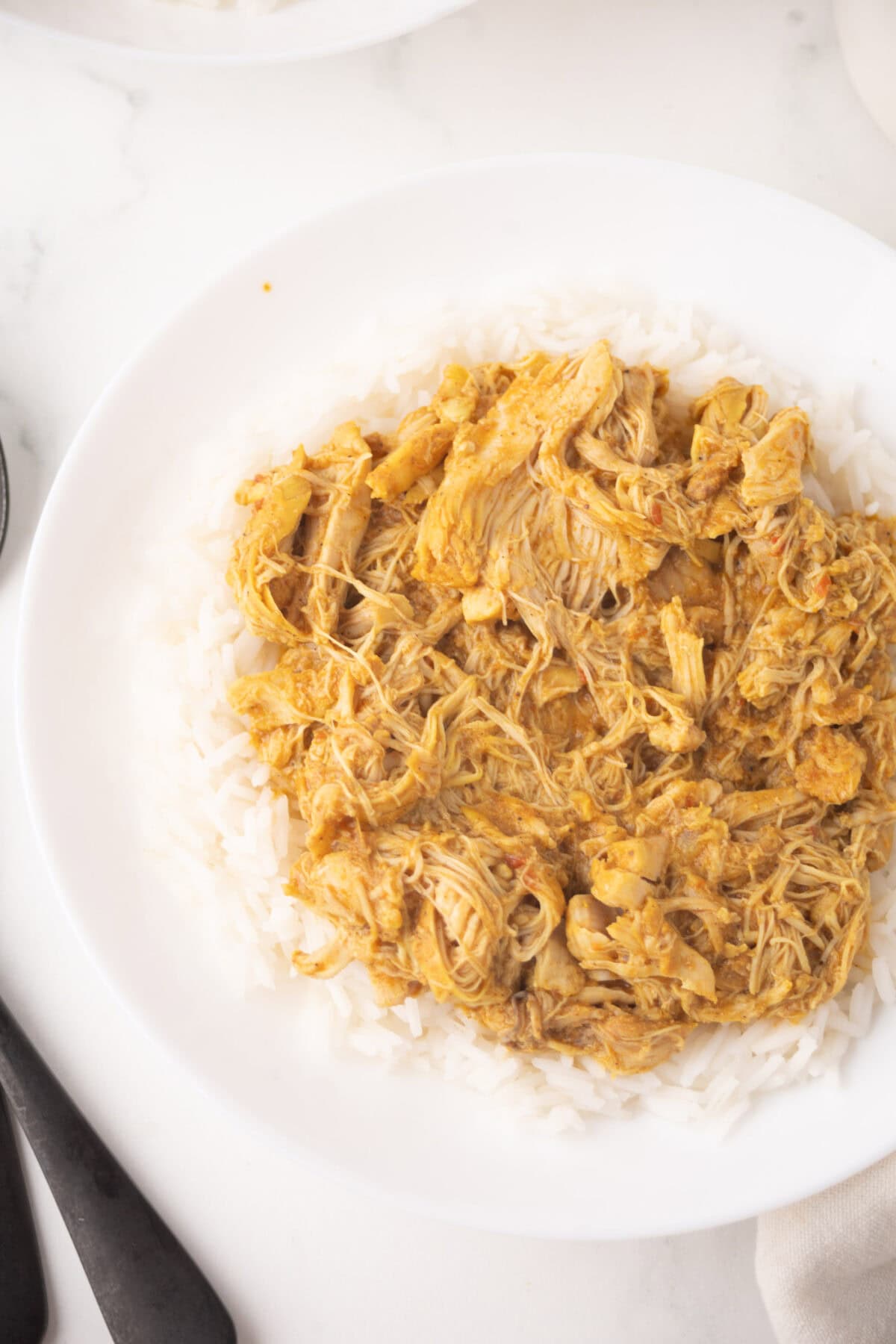 A white plate filled with shredded chicken korma served over a bed of white rice, with a fork and spoon placed beside the plate on a white marble surface.