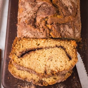 A loaf of cinnamon bread on a dark cutting board, with one slice cut to reveal a cinnamon sugar layer inside. Crumbs and cinnamon sugar are scattered around the board.