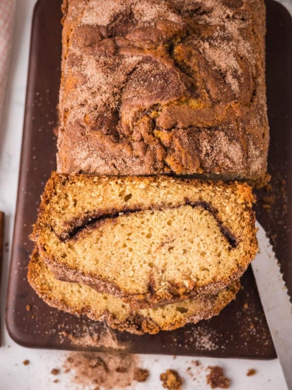 A loaf of cinnamon bread on a dark cutting board, with one slice cut to reveal a cinnamon sugar layer inside. Crumbs and cinnamon sugar are scattered around the board.