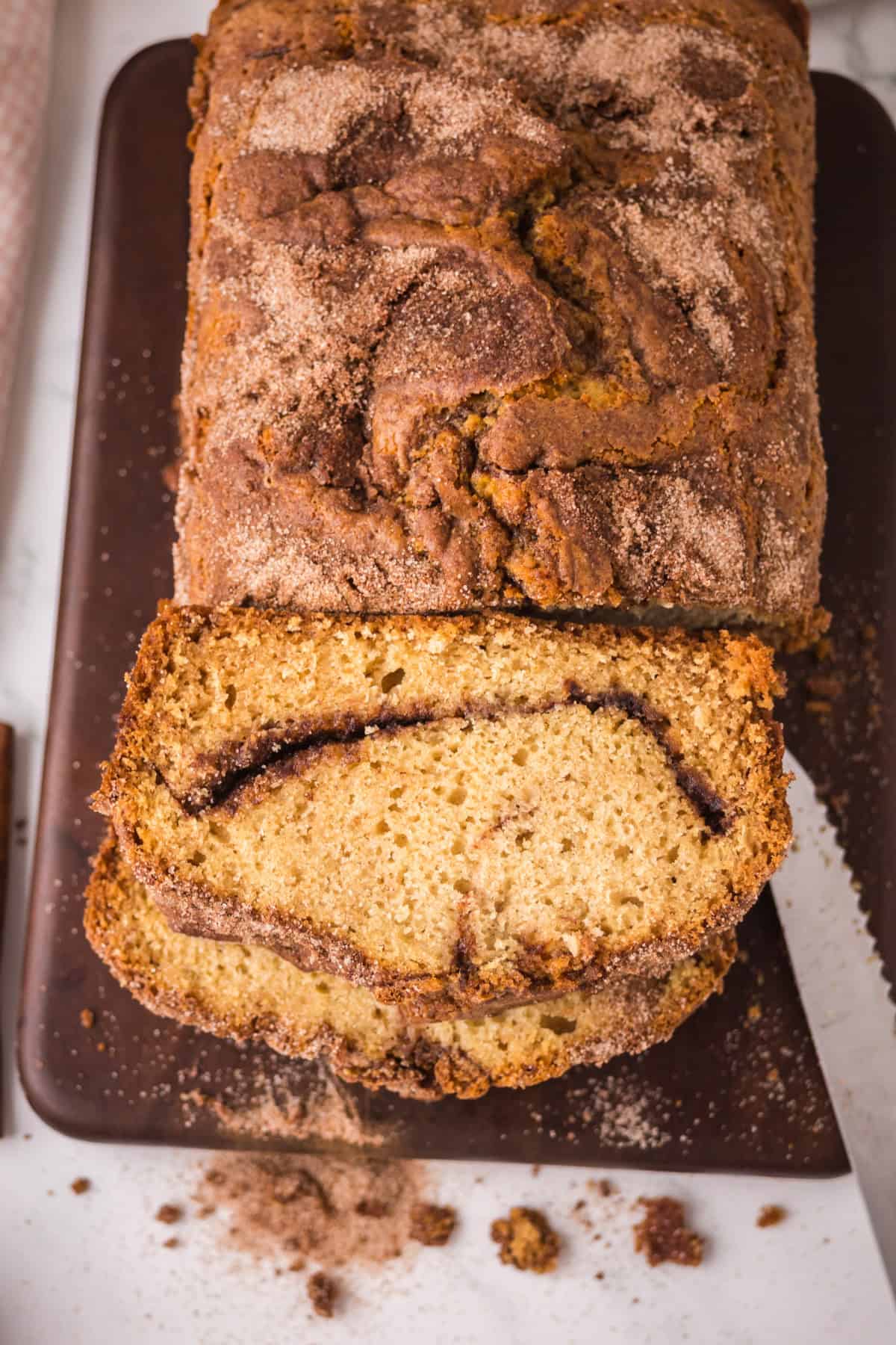 A loaf of cinnamon bread on a dark cutting board, with one slice cut to reveal a cinnamon sugar layer inside. Crumbs and cinnamon sugar are scattered around the board.