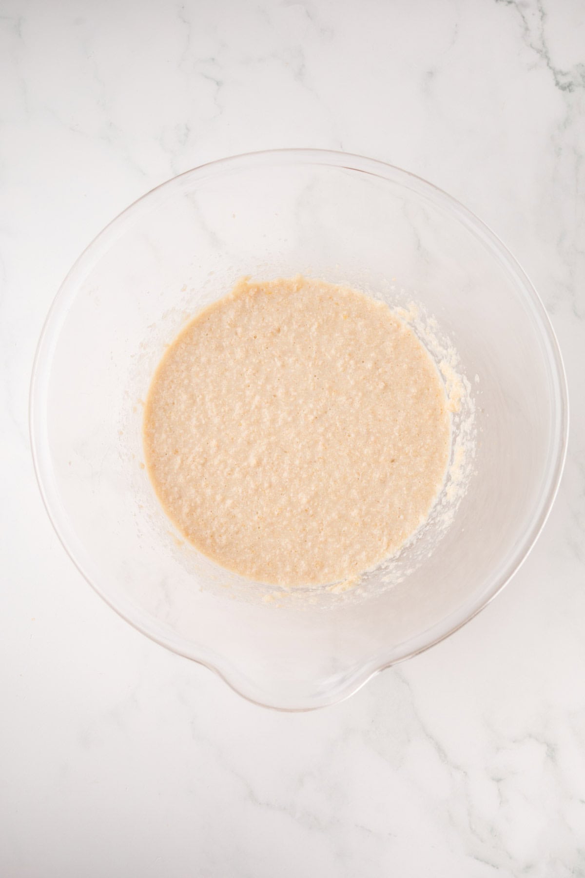 A glass mixing bowl containing a beige, slightly lumpy batter sits on a white marble countertop.