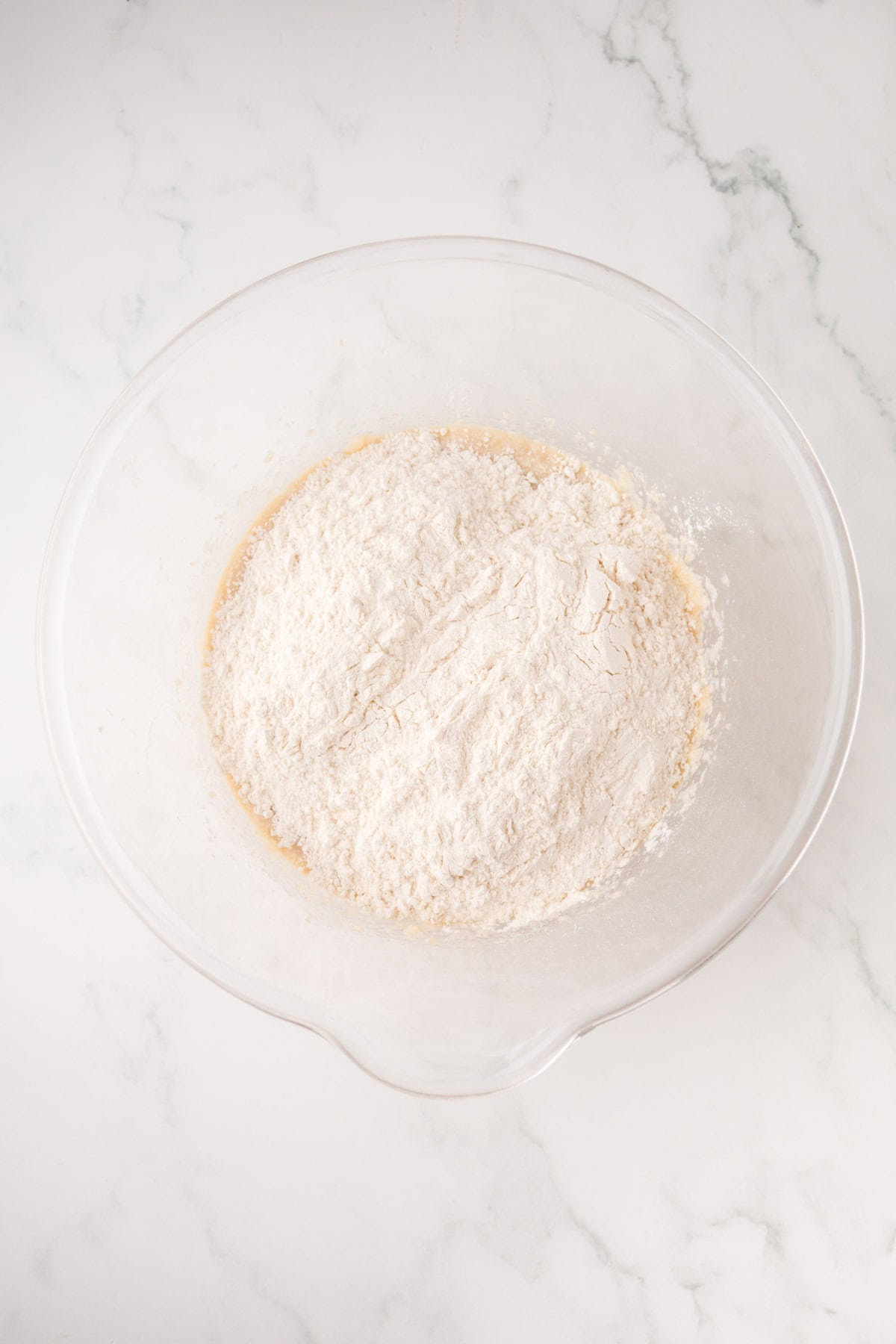 A clear glass mixing bowl containing flour on top of a beige mixture, placed on a white marble surface.