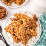 A plate of crockpot garlic honey chicken, shredded and saucy, served over white rice and topped with sesame seeds. A green checkered cloth, utensils, and small bowls of spices sit nearby, with a second plate in the background.