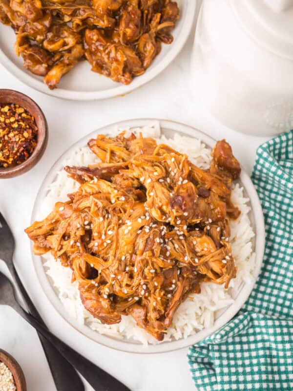 A plate of crockpot garlic honey chicken, shredded and saucy, served over white rice and topped with sesame seeds. A green checkered cloth, utensils, and small bowls of spices sit nearby, with a second plate in the background.