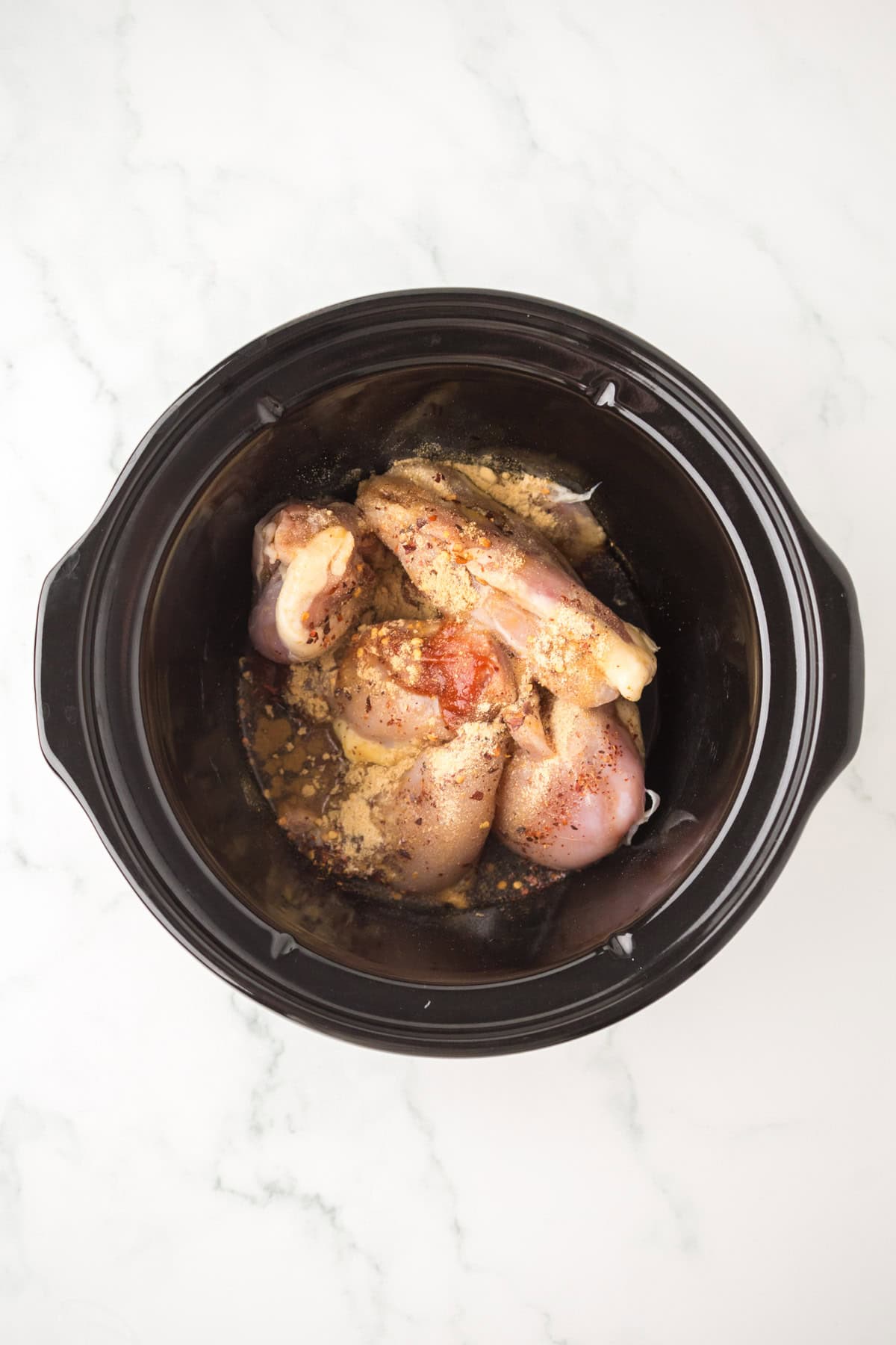 Raw chicken drumsticks with spices, garlic, and honey sauce are placed inside a black crockpot on a white marble surface, ready to be cooked.