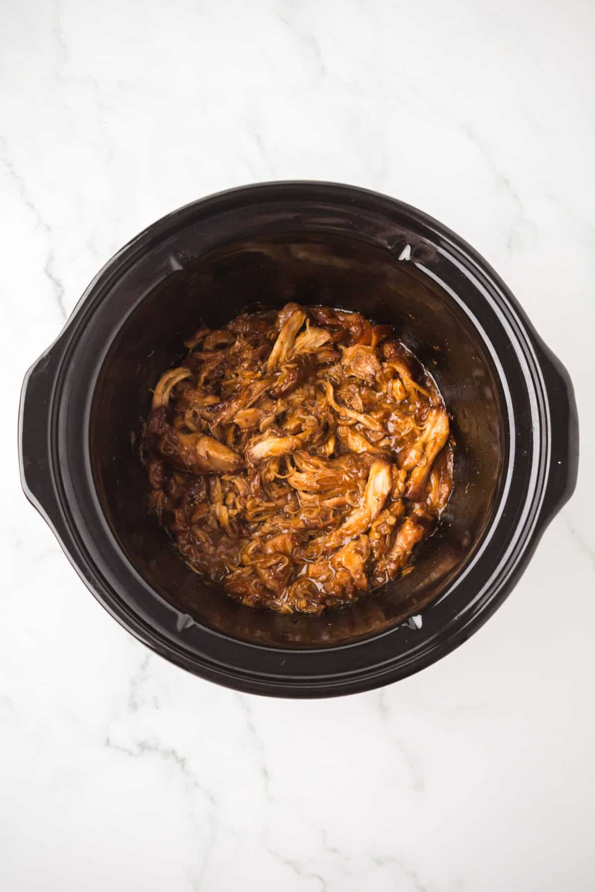 A black crockpot filled with shredded garlic honey chicken in a rich, dark sauce, placed on a white marble surface.