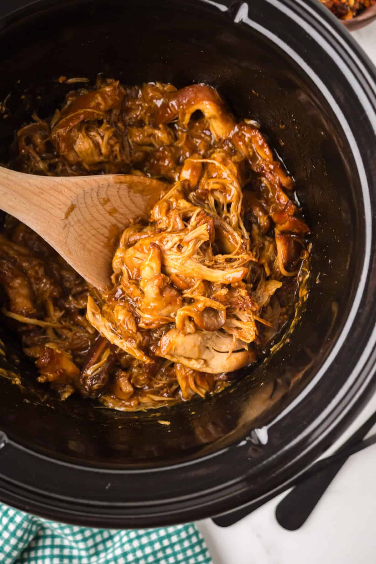 Shredded crockpot garlic honey chicken being stirred with a wooden spoon inside a black slow cooker, with a green gingham cloth partially visible on the counter nearby.