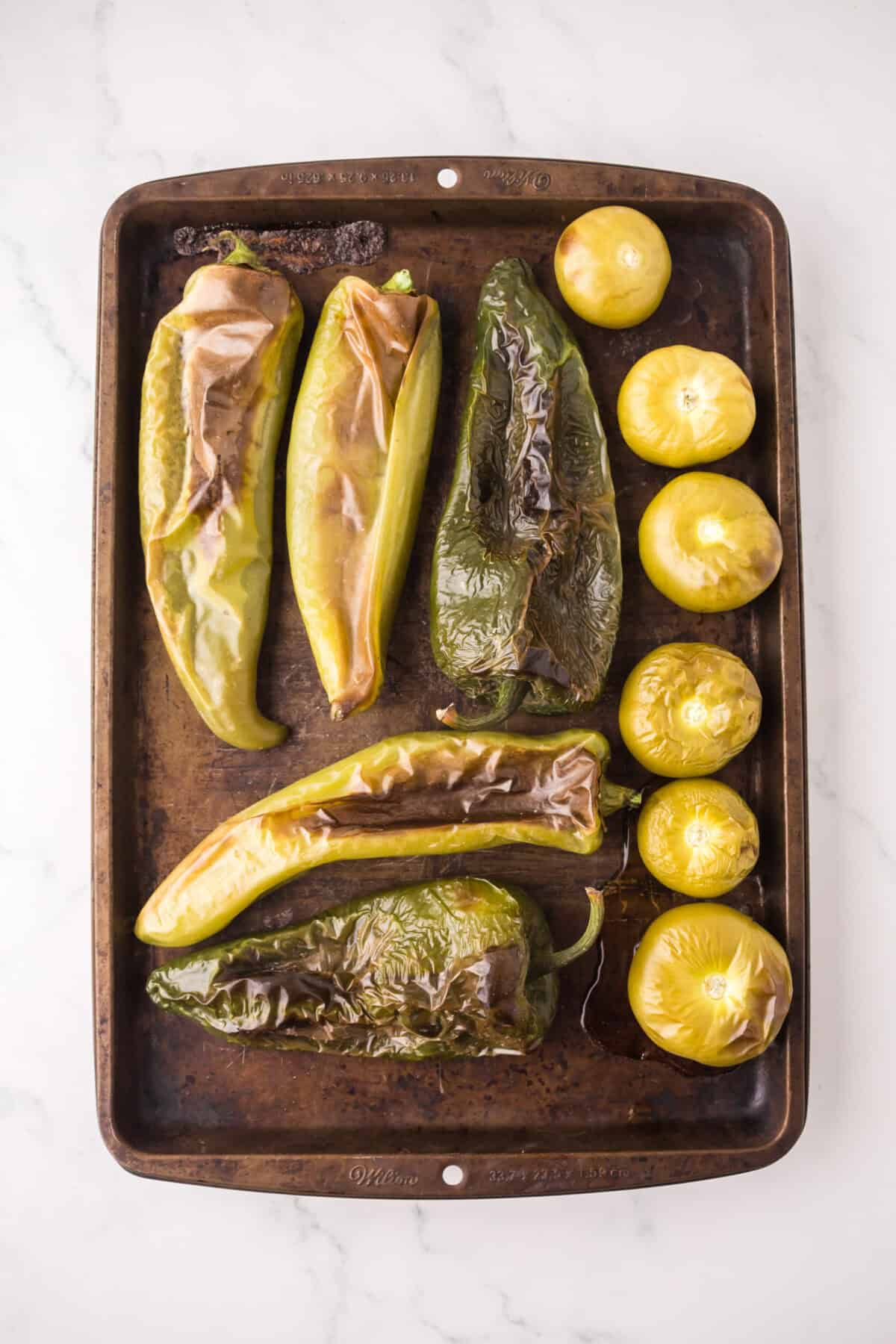 A baking sheet with roasted green peppers and yellow tomatoes, essential for a flavorful pork chili verde, rests on a white marble surface. The peppers and tomatoes have blistered, browned skins.